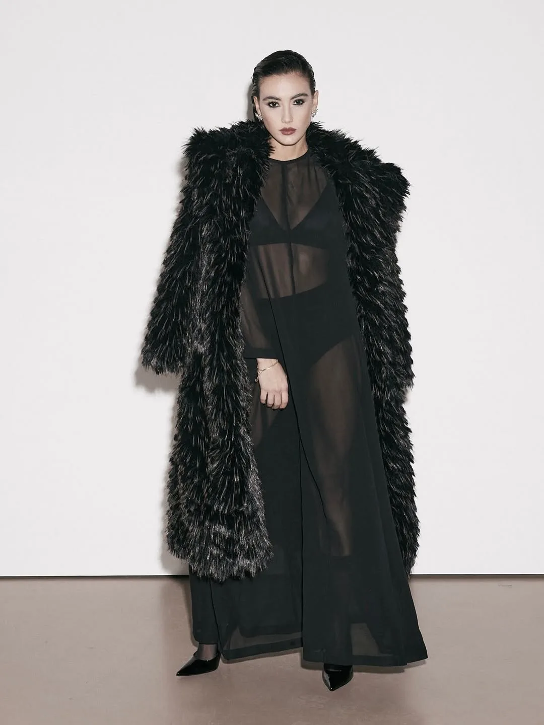 Zeudi standing in a long textured black coat in a bright studio.