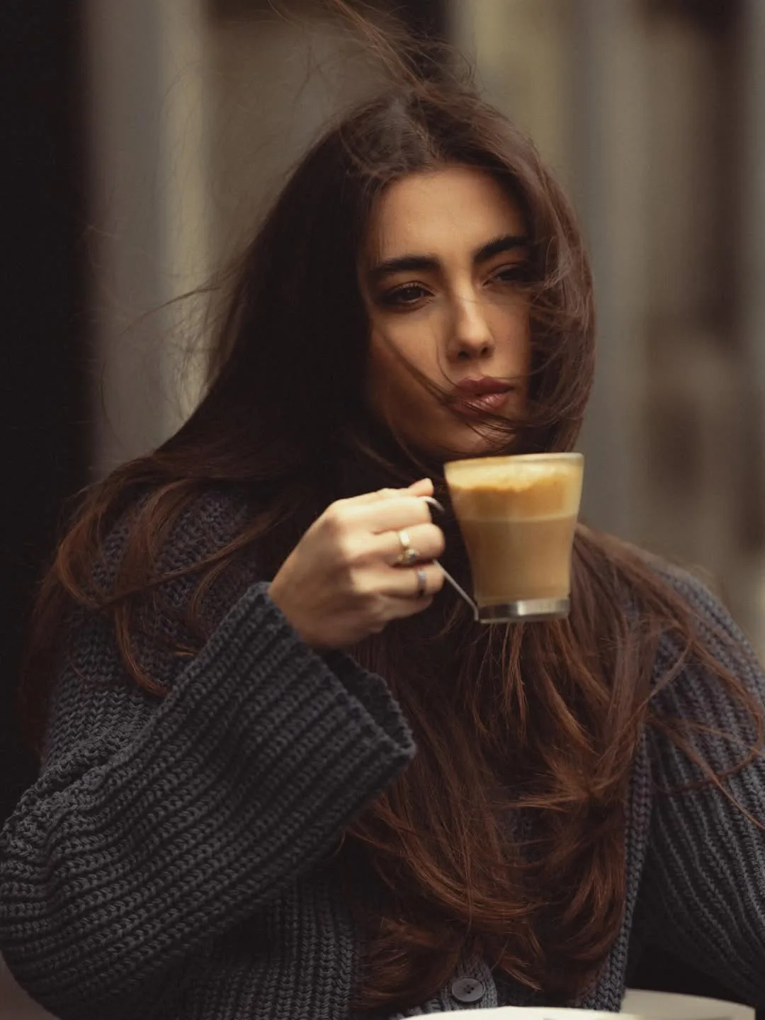 Zeudi holding a coffee cup near her face in a dark knitted sweater.