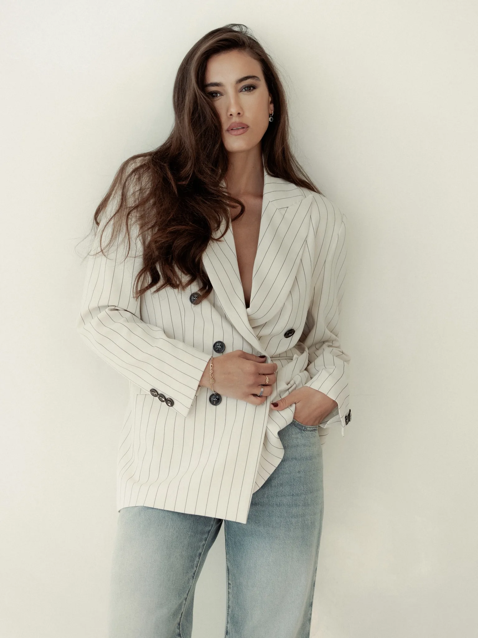 Zeudi standing against a pale wall in an oversized pinstriped blazer and jeans.