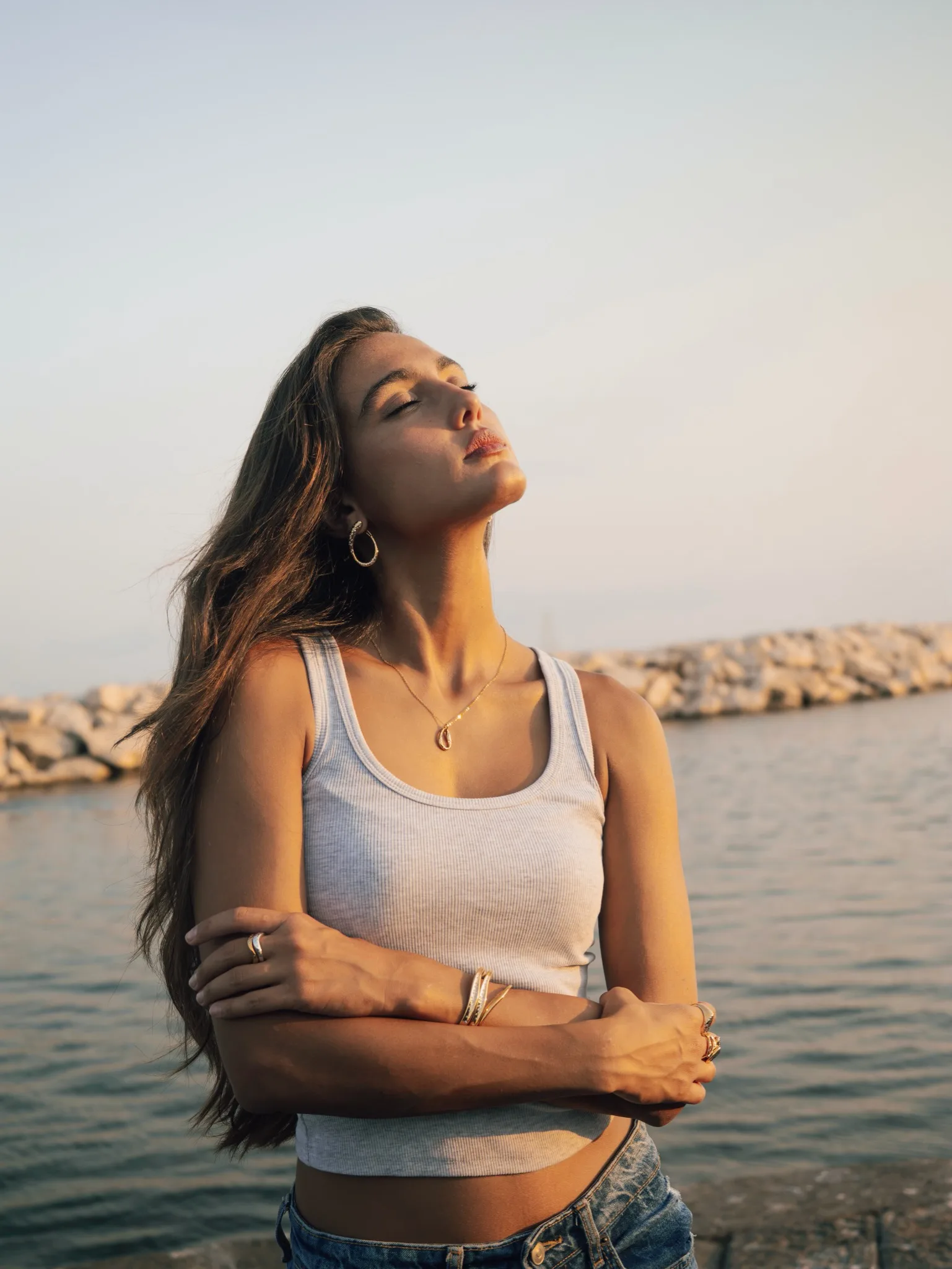 Zeudi standing beside the sea with her arms crossed in warm evening light.